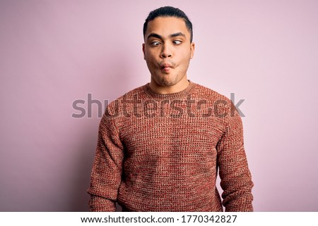 Young brazilian man wearing casual sweater standing over isolated pink background making fish face with lips, crazy and comical gesture. Funny expression.