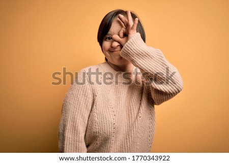 Young down syndrome woman wearing casual sweater over yellow background doing ok gesture with hand smiling, eye looking through fingers with happy face.