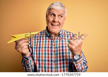 Senior handsome hoary man holding paper airplane standing over isolated yellow background pointing and showing with thumb up to the side with happy face smiling