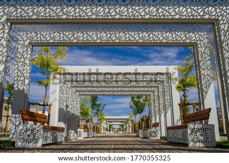 Alley in ethnic style. Openwork decor in ethnic style. Benches in the alley. Stylish white decorative elements in an urban environment. Central alley of Turkestan on a sunny day. Blue sky. Greenery