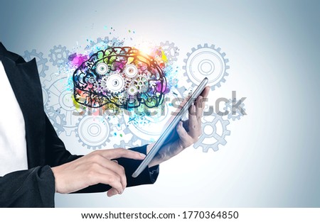 Hands of businesswoman using tablet computer near gray wall with colorful brain sketch drawn on it. Concept of brainstorming and artificial intelligence