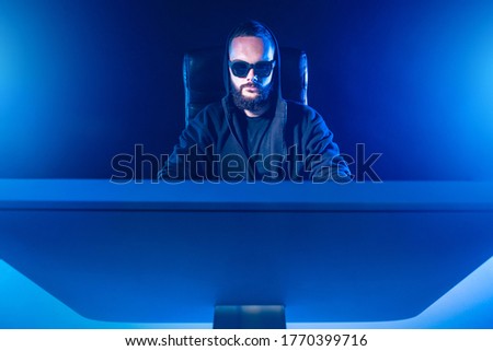 The concept of blogging. A man in black glasses and a hoodie behind a monitor. Blogger on a blue background. Place for text. Working at the computer. Creative photo with a blogger.