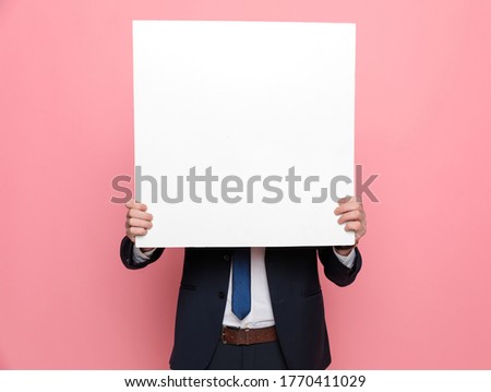 happy elegant businessman in suit hiding behind empty board and standing on pink background