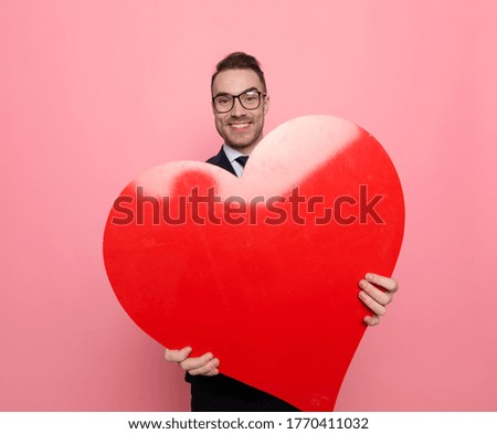 happy businessman in suit holding big red heart and smiling, standing on pink background
