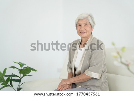 Smiling senior woman sitting on sofa and looking at camera. Awaken old woman with grey hair and pajamas in the early morning light. Portrait of elderly woman lying and smiling.