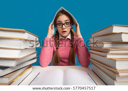 A lot of homework. Frightened and shocked teenager girl sits at table with stacks of books and covers her head with notebook, studio shot