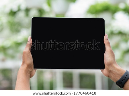 Industrial cultivation of potted plants with modern technology. Hands of african american girl with smartwatch holding tablet with empty screen on greenhouse background, pov, close up