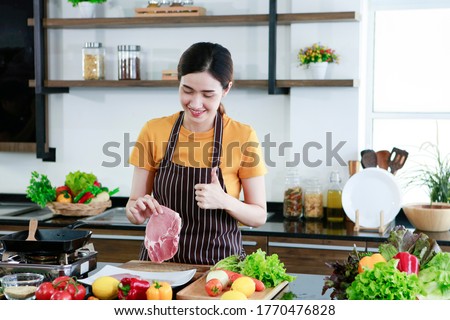 Asian women present fresh food. Shot housewife showing thumb up trying to cooking and showing pork steak in kitchen in house to take picture for sell food box online. Work from home / covid19 concept 