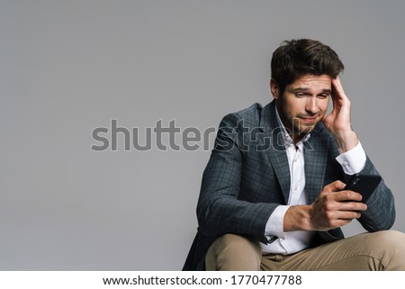 Photo of respectable confused businessman using cellphone while siting isolated over grey wall