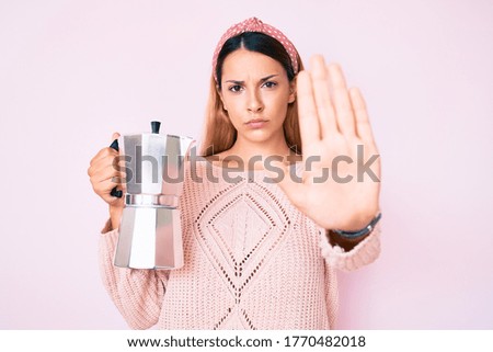 Young brunette woman holding italian coffee maker with open hand doing stop sign with serious and confident expression, defense gesture 