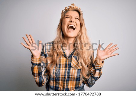 Young beautiful blonde woman wearing golden crown of queen over isolated white background celebrating mad and crazy for success with arms raised and closed eyes screaming excited. Winner concept