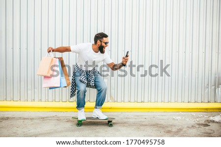 Fashionable hipster guy rides skateboard along an urban wall , wearing white t shirt , blue jeans and holding blank craft paper bags. Discount, sale, season sales.