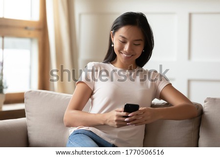 Smiling young vietnamese mixed race woman resting on cozy sofa, spending time online, chatting with friends in social network, using shopping mobile application, playing video game alone at home.