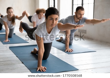Concentrated young female indian trainer showing bird dog pose at group yoga class. Fit barefoot diverse people practicing body balancing exercise in Parsva Balasana position on mat indoors in gym. Royalty-Free Stock Photo #1770508568