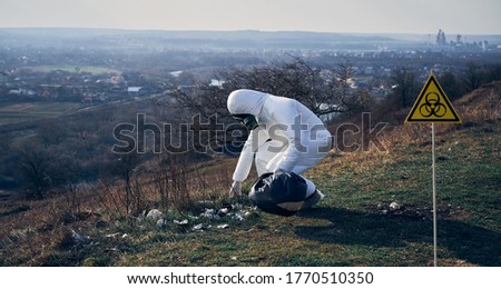 Environmentalist in protective suit and gas mask picking up garbage in abandoned grassy field, with biohazard sign. Concept of ecology, environmental pollution and biological hazard.