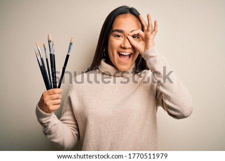Young beautiful artist asian woman holding paint brushes standing over white background with happy face smiling doing ok sign with hand on eye looking through fingers