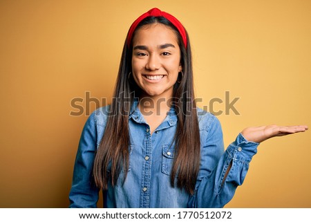 Young beautiful asian woman wearing casual denim shirt and diadem over yellow background smiling cheerful presenting and pointing with palm of hand looking at the camera.