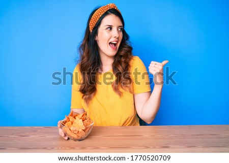 Young beautiful brunette woman sitting on the table eating nachos potato chips pointing thumb up to the side smiling happy with open mouth 