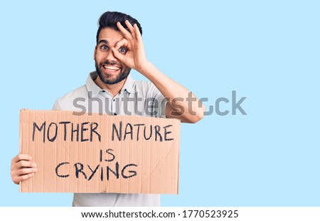 Young handsome man with beard holding mother nature is crying cardboard banner smiling happy doing ok sign with hand on eye looking through fingers 