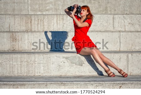 Young beautiful girl photographer in a bright red dress focuses on a model with a large digital camera