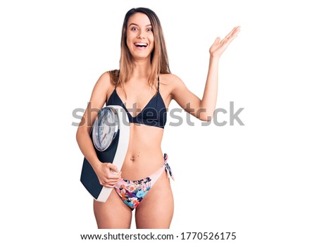 Young beautiful girl wearing bikini holding weighing machine celebrating victory with happy smile and winner expression with raised hands 