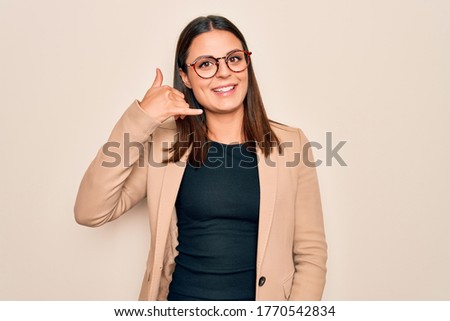 Young beautiful brunette businesswoman wearing jacket and glasses over white background smiling doing phone gesture with hand and fingers like talking on the telephone. Communicating concepts.