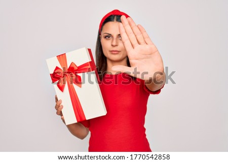 Young beautiful brunette woman holding birthday gift over isolated white background with open hand doing stop sign with serious and confident expression, defense gesture