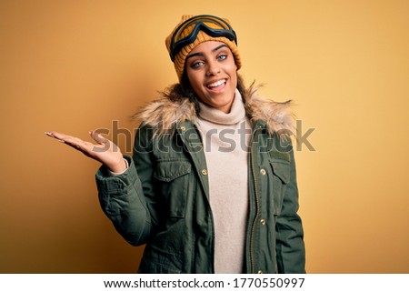 Young african american afro skier girl wearing snow sportswear and ski goggles smiling cheerful presenting and pointing with palm of hand looking at the camera.