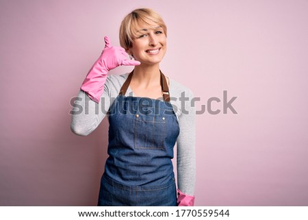 Young blonde cleaner woman with short hair wearing apron and gloves over pink background smiling doing phone gesture with hand and fingers like talking on the telephone. Communicating concepts.