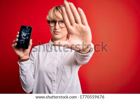 Young blonde woman with short hair holding broken smartphone over red background with open hand doing stop sign with serious and confident expression, defense gesture