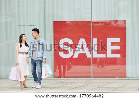 Young happy Vietnamese couple with shopping bags standing at big sale banner outside the shopping mall