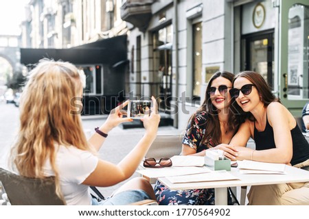 Three beautiful friends met at a cafe on the summer terrace. Attractive young women sit at a table outdoor, one of them takes pictures of the other two on the phone