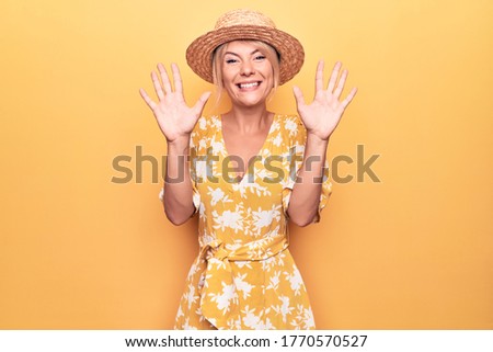 Beautiful blonde woman on vacation wearing summer hat and dress over yellow background showing and pointing up with fingers number ten while smiling confident and happy.