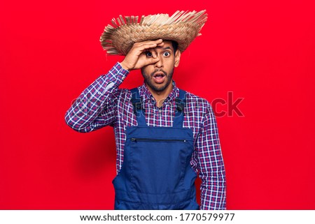 Young latin man wearing farmer hat and apron doing ok gesture shocked with surprised face, eye looking through fingers. unbelieving expression. 