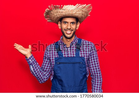 Young latin man wearing farmer hat and apron smiling cheerful presenting and pointing with palm of hand looking at the camera. 