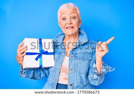 Senior beautiful woman with blue eyes and grey hair holding gift smiling happy pointing with hand and finger to the side 