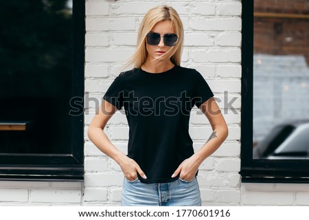 Stylish blonde girl wearing black t-shirt and glasses posing against street , urban clothing style. Street photography Royalty-Free Stock Photo #1770601916