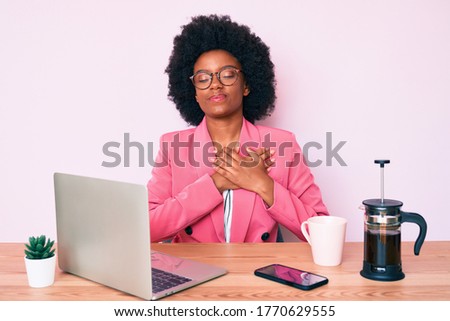 Young african american woman working at desk using computer laptop smiling with hands on chest, eyes closed with grateful gesture on face. health concept. 