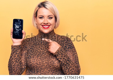 Young beautiful blonde plus size woman holding broken smartphone showing cracked screen smiling happy pointing with hand and finger