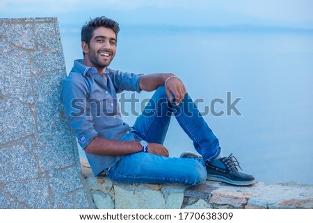 Man smiling looking at camera happy celebrating freedom while sitting on a concrete wall bridge above the sea. Positive person, peace mind concept. Free happy guy enjoying sunset