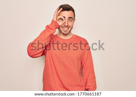 Young handsome man with blue eyes wearing casual sweater standing over white background doing ok gesture with hand smiling, eye looking through fingers with happy face.