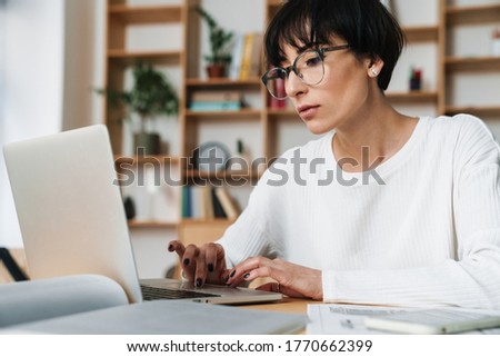 Photo of serious beautiful woman in eyeglasses working with laptop while sitting at table in office