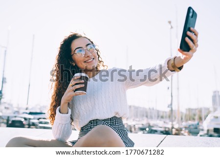 Happy Hispanic female user taking selfie pictures during coffee break in city, youthful millennial blogger smiling at front camera while clicking pictures via filter app on modern smartphone