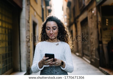 Backlit young content lady in eyeglasses and white sweater browsing smartphone while walking along narrow old street on sunny day
