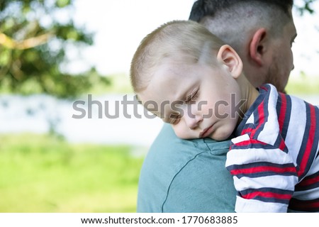 A young child is sleeping in his father's safe and protective embrace while on walk. Portrait of cute adorable blond caucasian toddler boy sleeping on fathers shoulder in park. Single father concept.