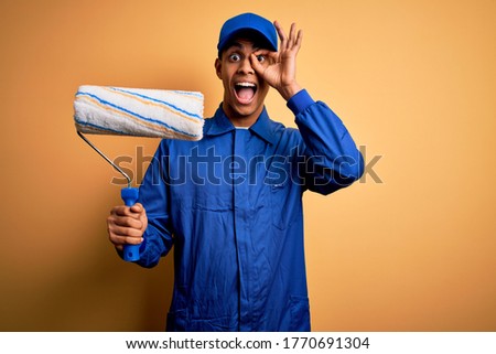 Young handsome african american painter man wearing uniform using painting roller with happy face smiling doing ok sign with hand on eye looking through fingers