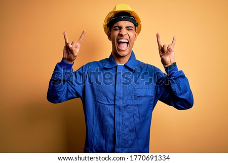 Young handsome african american worker man wearing blue uniform and security helmet shouting with crazy expression doing rock symbol with hands up. Music star. Heavy music concept.