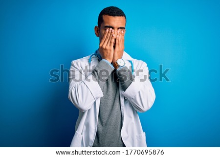 Handsome african american doctor man wearing coat and stethoscope over blue background rubbing eyes for fatigue and headache, sleepy and tired expression. Vision problem