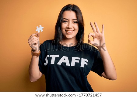 Young brunette worker woman wearing staff t-shirt as uniform showing puzzle piece as teamwork doing ok sign with fingers, excellent symbol