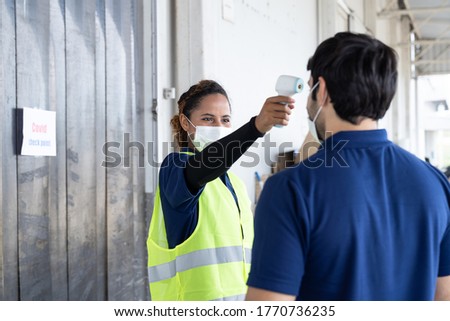 Woman worker wearing face mask using infrared forehead thermometer with man worker in front of the warehouse at the factory. Factory industry worker operating concept. Royalty-Free Stock Photo #1770736235